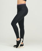 Leggings deportivos negros para mujer de silueta ajustada, talle alto con vivo morado y bota tubo. Vista lateral.